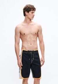 Černo-bílé plavky s žlutými detaily, vybavené stahovací šňůrkou v pase, bočními kapsami a sportovním střihem. Hladká textura.