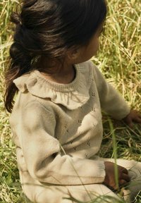 Pull en tricot beige avec un col volants, présentant un motif texturé. L'enfant est assis dans l'herbe, avec des cheveux sombres et lâches.