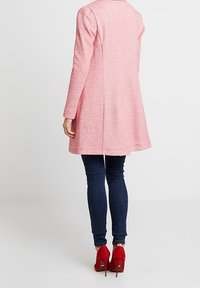 Cappotto rosa strutturato con maniche lunghe, lunghezza fino a metà coscia e cuciture posteriori. Indossato sopra jeans scuri e décolleté rossi, con un design pulito.