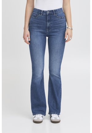 Donna che indossa jeans a vita alta, blu medio con fondo svasato e tasche frontali, e scarpe da ginnastica bianche, in piedi contro uno sfondo neutro.