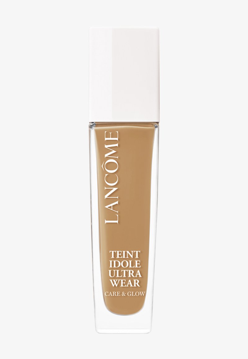 Botella de base de vidrio transparente con líquido beige en su interior y una tapa alta blanca. Presenta las etiquetas "Lancôme" y "Teint Idole Ultra Wear".