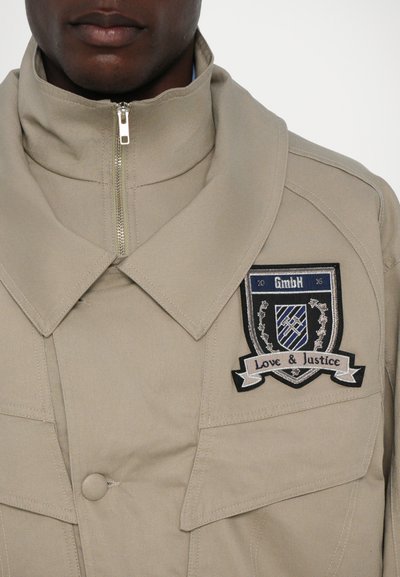 Beige Reißverschlussjacke mit hohem Kragen, ausgestattet mit einem blau-schwarzen gestickten Patch, der "GmbH" und "Love & Justice" auf der Vorderseite zeigt.