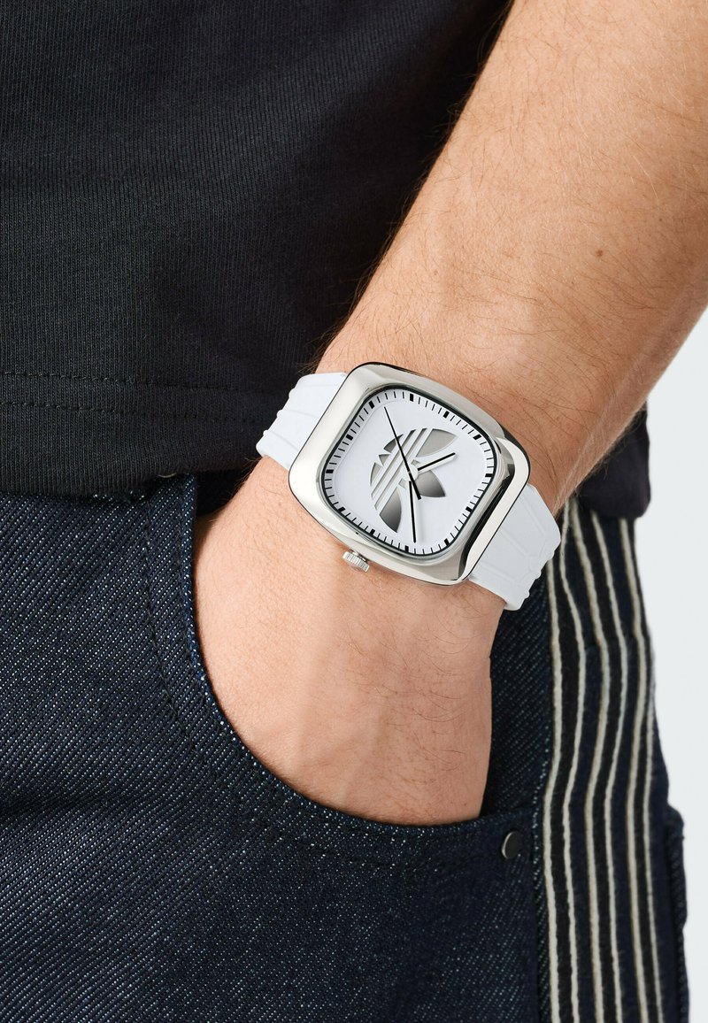 Orologio da polso con cassa quadrata argentata, cinturino bianco e design unico del logo sul quadrante, indossato al polso di una persona con la mano nella tasca dei jeans.