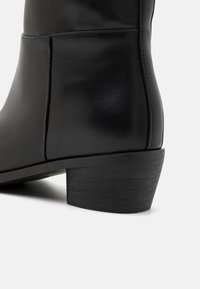 Černá kožená kotníková bota s kulatou špičkou, jemnými švy a texturovaným černým nízkým podpatkem. Hladký povrch a elegantní design.