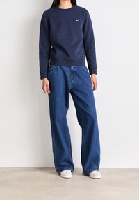 Sudadera navy con cuello redondo y parche de logo, combinada con jeans azules holgados y zapatillas blancas, de pie frente a un fondo neutro.
