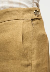 Κοντινή λήψη της μέσης μπεζ λινών παντελονιών με μεγάλο κουμπί και πλευρικό φερμουάρ, σε άτομο που φοράει ανοιχτόχρωμο τοπ.