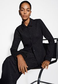 Blazer nero scintillante in maglia con colletto, chiusura con bottone e maniche lunghe, abbinato a pantaloni coordinati. Tessuto liscio e design aderente.