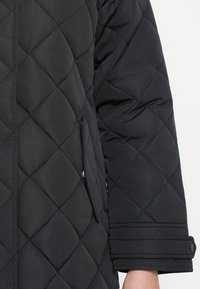 Quilted black jacket featuring a diagonal pattern, a side pocket with flap, and buttoned cuffs. Soft texture and mid-length sleeves.
