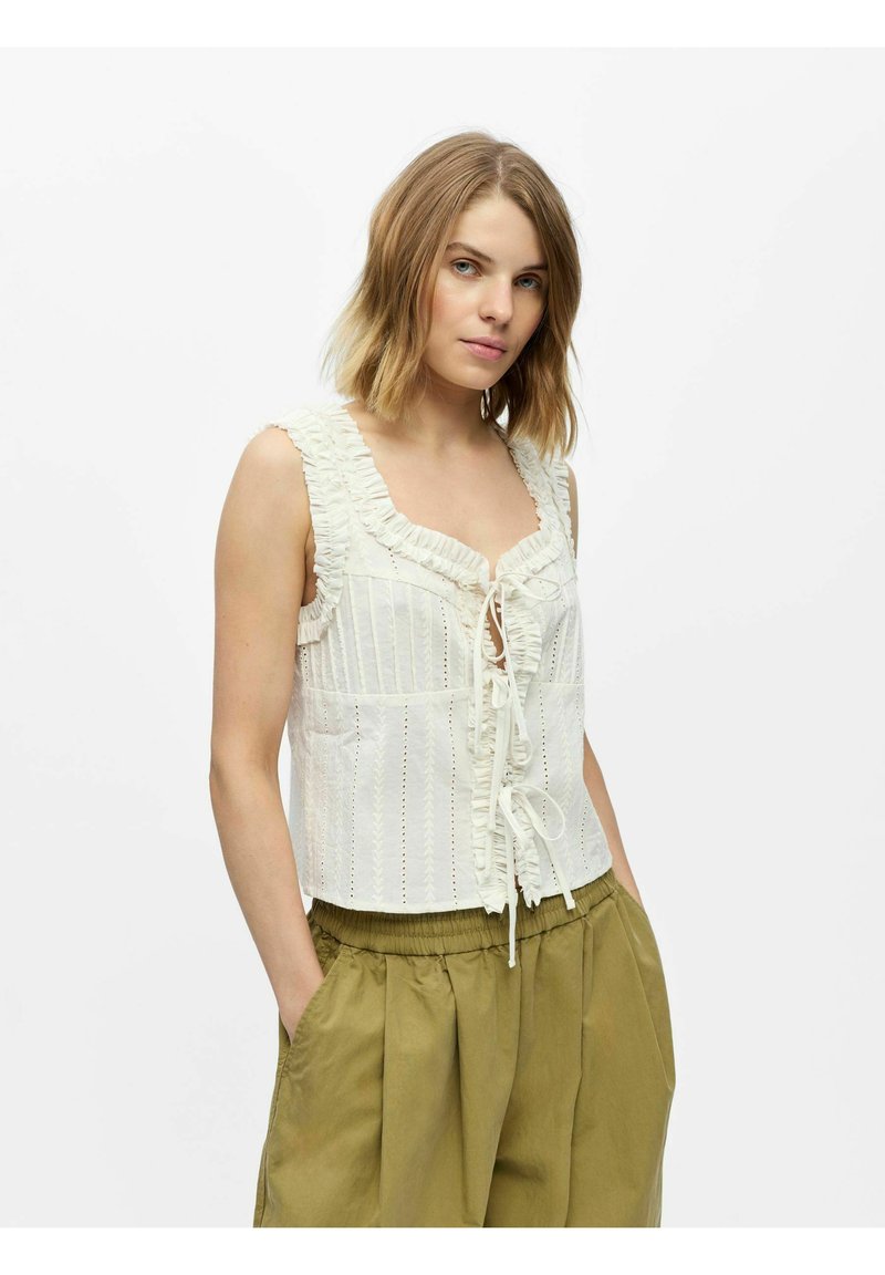 Jeune femme aux cheveux blonds mi-longs portant un haut blanc sans manches avec des volants et un pantalon ample vert olive, les mains dans les poches.