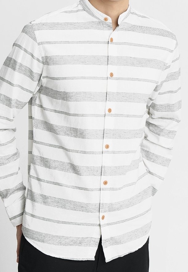 Homme portant une chemise blanche à manches longues avec des rayures horizontales gris clair et des boutons en bois, debout, les mains dans les poches.