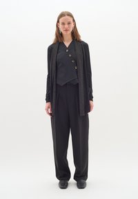Svart, texturerad cardigan över en svart topp med guldknappar, vida svarta byxor och svarta skor. Enkel, elegant design med minimala accenter.