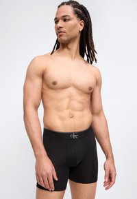 Sportlicher Mann mit langen Dreadlocks, der schwarze Calvin Klein Boxershorts trägt, vor schlichtem hellem Hintergrund stehend und zur Seite blickend.