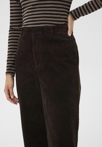 Pantalon en velours côtelé marron foncé avec une texture à côtes verticales, doté d'une ceinture avec passants et de poches latérales. Main reposant sur le côté.