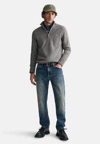 Grauer Fleece-Half-Zip-Pullover, darunter ein blaues Hemd mit Kragen, blaue Jeans, schwarzer Gürtel und schwarze Schnürschuhe. Grüner Hut mit dem Buchstaben "G".