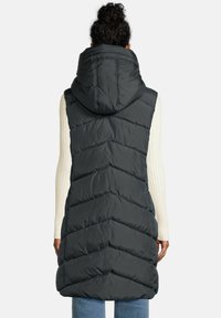Vrouw in een zwarte doorgestikte mouwloze capuchonvest over een witte geribbelde longsleeve top en blauwe jeans, gezien van achteren.