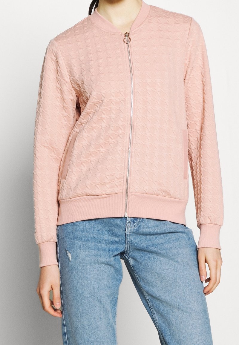 Donna che indossa una giacca zip-up di un rosa chiaro con texture, maniche e orlo a costine, abbinata a jeans blu chiaro strappati.