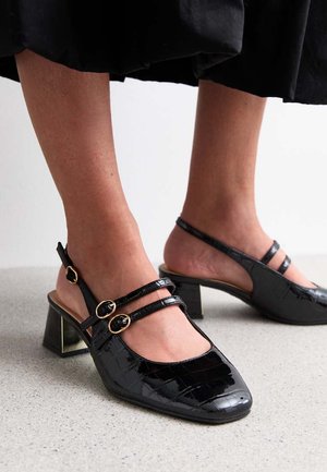 Zwarte gelakte leren slingback hakken met vierkante neuzen, twee dunne banden met gouden gespen, en blokhakken met een gestructureerd oppervlak.