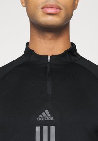 Schwarzes sportliches Zip-up-Oberteil mit hohem Kragen, versehen mit einem silbernen Adidas-Logo und einem Design aus drei horizontalen Streifen auf der Vorderseite. Glatte Textur.