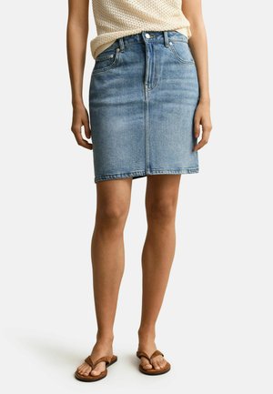 Femme portant une jupe en denim bleu clair, un haut en maille beige et des sandales marron, debout devant un fond blanc uni.