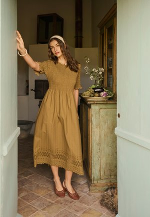 Žena v hořčicovozlatej midi šatách a s čelenkou stojí v dverách rustikálnej kuchyne s drevenou skriňou a miskou zeleniny.