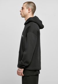 Mann mit kurzen Haaren, der einen schwarzen Hoodie und schwarze Hosen trägt, im Profil vor einem einfarbigen hellgrauen Hintergrund stehend.