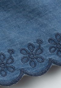 Tissu en denim avec des bords festonnés et une broderie florale bleu marine le long du bas. La texture est lisse avec des motifs de tissage visibles.