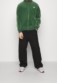 Groene velvet jas met ritssluiting, witte strepen aan de zijkant en Nike-logo, gecombineerd met losse zwarte broek en witte sneakers met rode accenten.