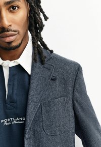 Navy-Blazer im Fischgrätenmuster mit strukturiertem Finish und Reverskragen, kombiniert mit einem dunklen, gerippten Hemd mit Kragen, das weiße Akzente aufweist.