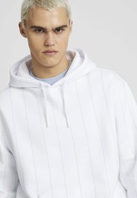Hoodie blanc avec des rayures verticales bleu clair, cordons de serrage et une poche à l'avant. Tissu doux avec une coupe décontractée, superposé sur une chemise bleue.