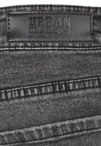 Le tissu denim noir présente une surface texturée avec des coutures distinctes. Une patch en cuir noir, embossé avec "URBAN CLASSICS DENIM FACTORY", est attaché.