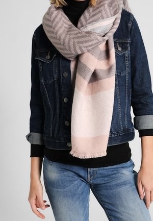 Personne portant une veste en denim bleu, un col roulé noir, un jean bleu clair, et une grande écharpe à motifs roses et gris enroulée autour du cou.