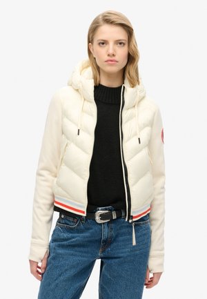 Veste blanche ronde avec une capuche, des poignets côtelés et une fermeture éclair. Présente un ourlet bicolore avec des bandes rouges, bleues et blanches. Portée sur un pull noir.