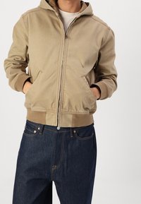 Tan zip-up hoodie with a hood, ribbed cuffs, and side pockets, paired with dark denim jeans. Visible stitching and a relaxed fit.