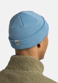 Light blue knitted beanie with a folded cuff and a small logo patch at the back, worn with a beige fleece jacket.