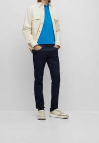 Vit bomullskjorta med jacka, blå polo, mörka denimjeans, beige och krämfärgade sneakers. Jackan har två bröstfickor och en avslappnad passform.