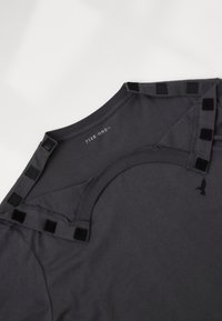 Σκούρο γκρι μπλούζα με στρογγυλή λαιμόκοψη, μαλακό ύφασμα και χαρακτηριστικές μαύρες ταινίες βέλκρο στους ώμους και ένα λογότυπο κοντά στο στρίφωμα.