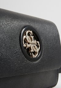 Sort tekstureret læder håndtaske med et cirkulært logo i goldmetal, med en elegant klaplukning og glatte kanter.