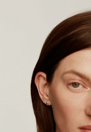 Primer plano de la oreja derecha de una mujer con cabello castaño corto recogido detrás, llevando un pequeño pendiente de aro de oro con tachuelas en forma de pirámide.