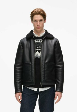 Joven con una chaqueta negra de cuero con cuello de borrego sobre un suéter estampado y camisa blanca, de pie con expresión neutral.