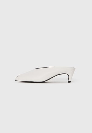 Sapato mule branco de bico pontiagudo com um salto curvo baixo e sola preta sobre um fundo branco liso.