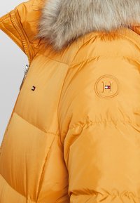 Orange puffjacka med en faux fur-krage, med ett logopatch och en elegant dragkedjestängning. Tyget har en slät, glänsande textur.
