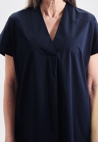 Marinblå klänning med V-formad halsringning och korta ärmar, tillverkad av mjukt tyg. Har en avslappnad passform och subtila veck framtill.