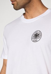 Λευκό βαμβακερό μπλουζάκι με στρογγυλή λαιμόκοψη και μαύρο κυκλικό λογότυπο με αετό και κείμενο «Deutscher Fussball Bund» στο αριστερό στήθος.