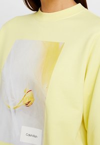 Sudadera amarilla con un estampado brillante de una flor, cuello redondo y un logo de Calvin Klein en la parte inferior delantera. Material de algodón suave.