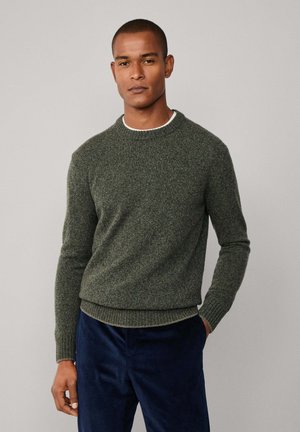 Man standing with one hand in pocket wearing an olive green sweater over a white shirt and navy blue pants against a plain background.