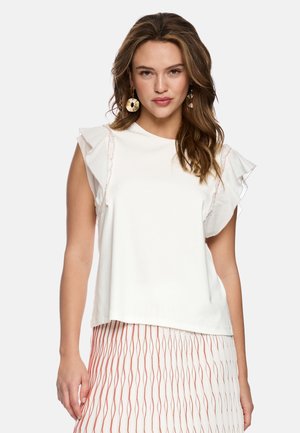 Frau mit welligem Haar, die goldene runde Ohrringe, ein weißes ärmelloses Oberteil mit Rüschenschultern und einen weißen Rock mit vertikalen orangen Mustern trägt.