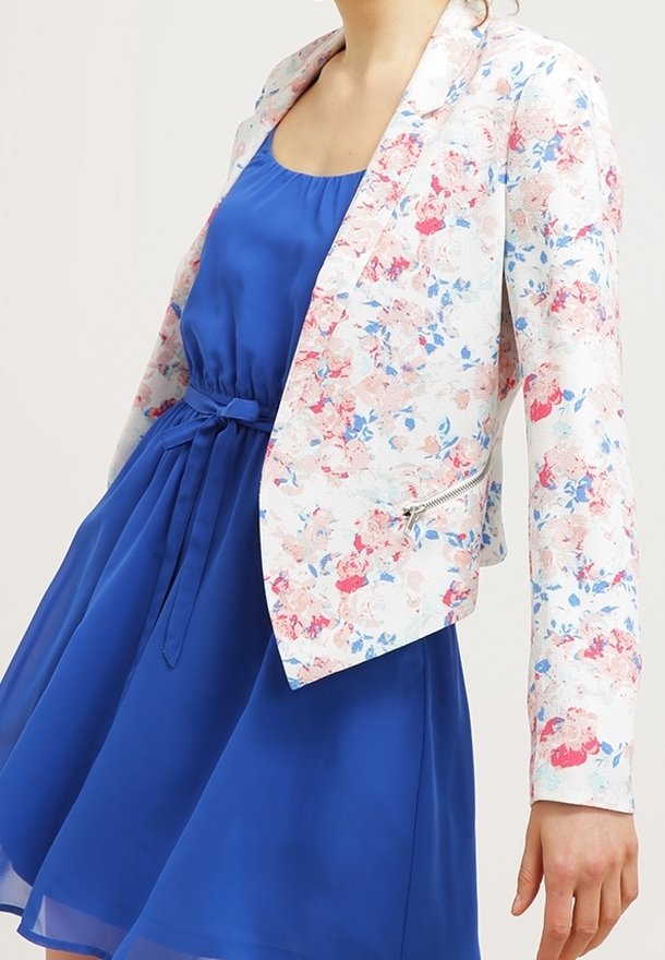 Femme portant une robe bleu roi sans manches avec une ceinture à la taille et un blazer blanc à motif floral avec des détails zippés.