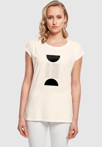 Krémfarvet t-shirt med korte ærmer, der har et sort geometrisk design med to halvcirkler og vandrette linjer, lavet af blødt bomulds materiale.