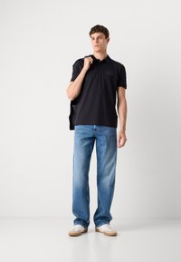 Camisa polo preta de mangas curtas com um logo discreto, combinada com jeans de ganga azul clara e ténis bege. Roupa casual com um corte moderno.