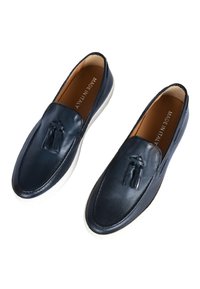 Scarpe blu in pelle senza lacci con design della punta cucito e un accento con nappina, dotate di suola in gomma bianca e rivestimento interno beige.